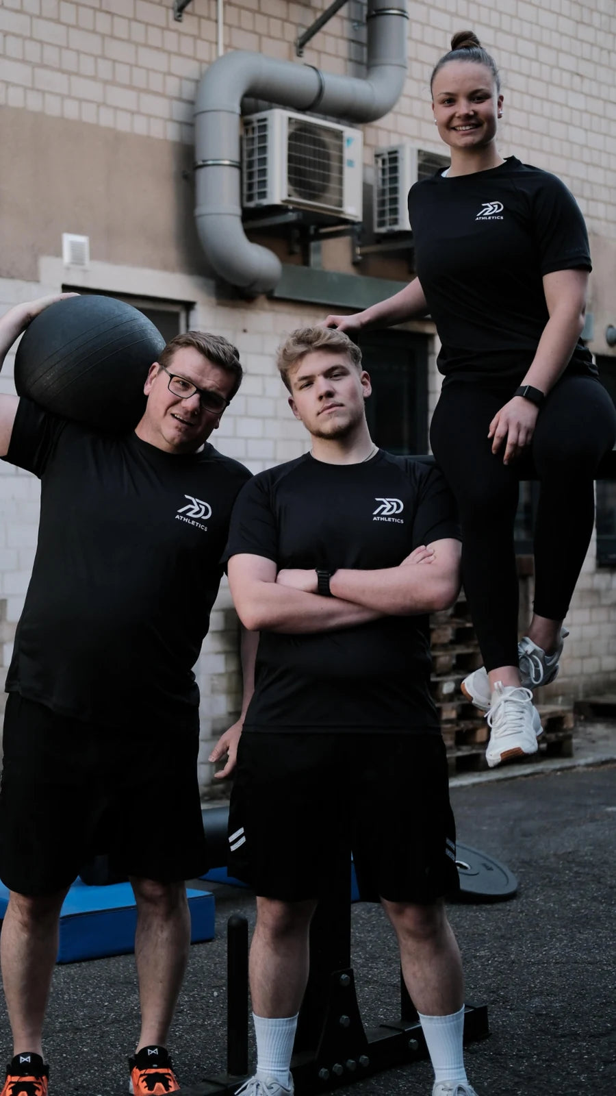 Drei Mitglieder des PGO-Athletics-Teams in schwarzen Trainingsshirts im Hinterhof-Gym, einer mit Medizinball auf der Schulter