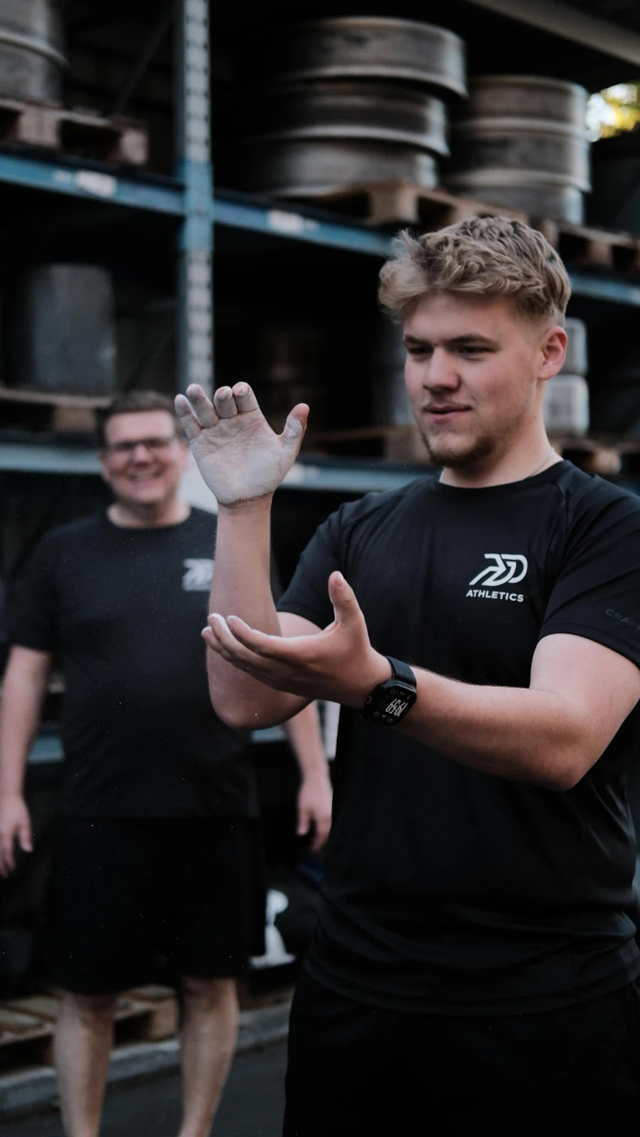 Sportler kreidet sich vor einem Regal voller Hantelscheiben die Hände ein, im Hintergrund ein lachender Trainingspartner