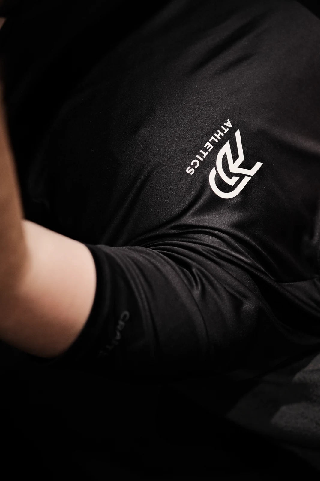 Nahaufnahme eines schwarzen PGO ATHLETICS Shirts mit weißem Logo-Print, getragen von einer Person mit angewinkeltem Arm